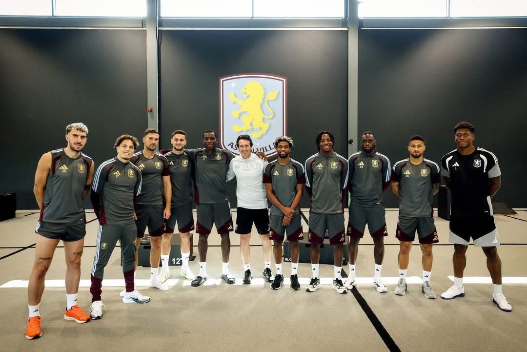 Aston Villa players and coaching staff standing beneath the club crest at the training ground