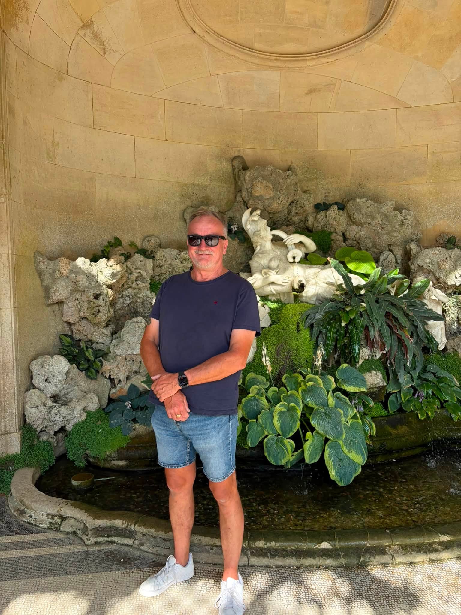 John standing beside a shaded water feature with stone figures and lush planting